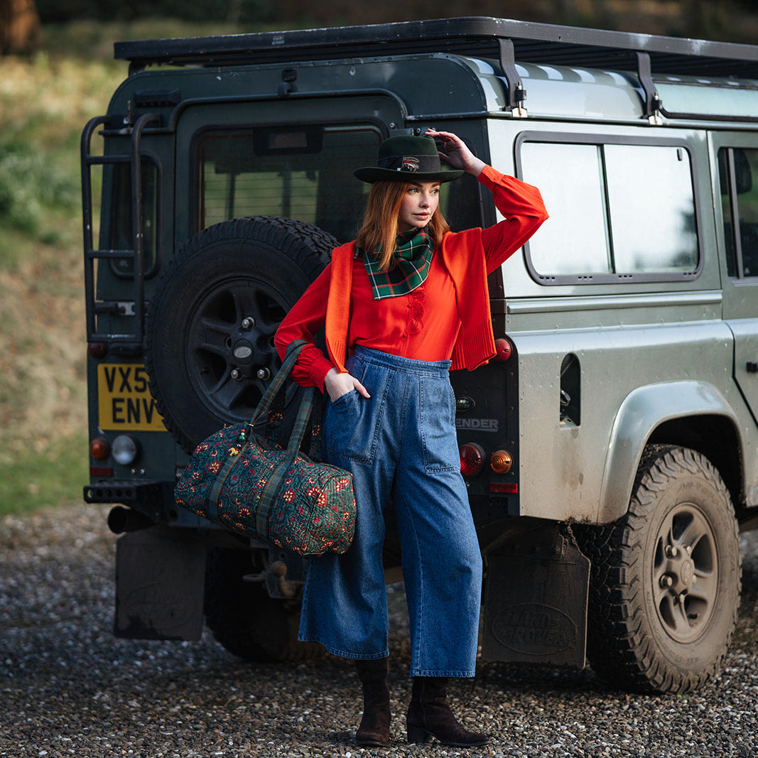 Woman with tartan scarf and blue jeans standing next to a Land Rover with a suitcase or weekend bag.